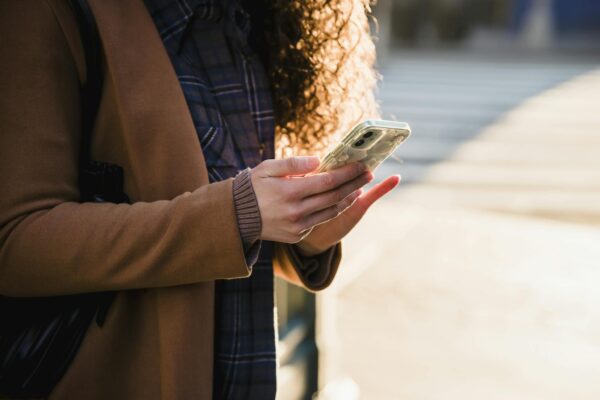 Crop anonymous female with curly hair in warm stylish coat using mobile phone on blurred background of urban street